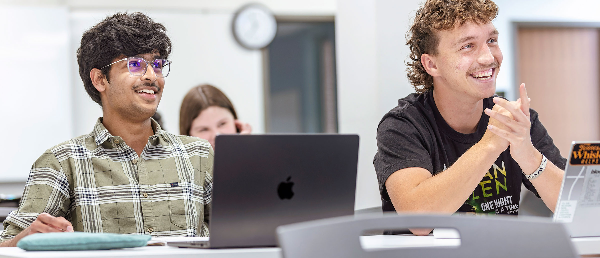 two male students sitting at desks look toward the front of a classroom while smiling